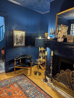 A room with a dark blue wall, a framed picture, a lamp, a candelabra, a chair, a rug, and a fireplace.