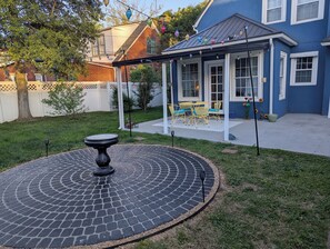 Eine Terrasse mit einem kreisförmigen Steinmuster, ein überdachter Sitzbereich mit gelben Stühlen und ein Haus mit blauen Wandverkleidungen und weißen Zierleisten.