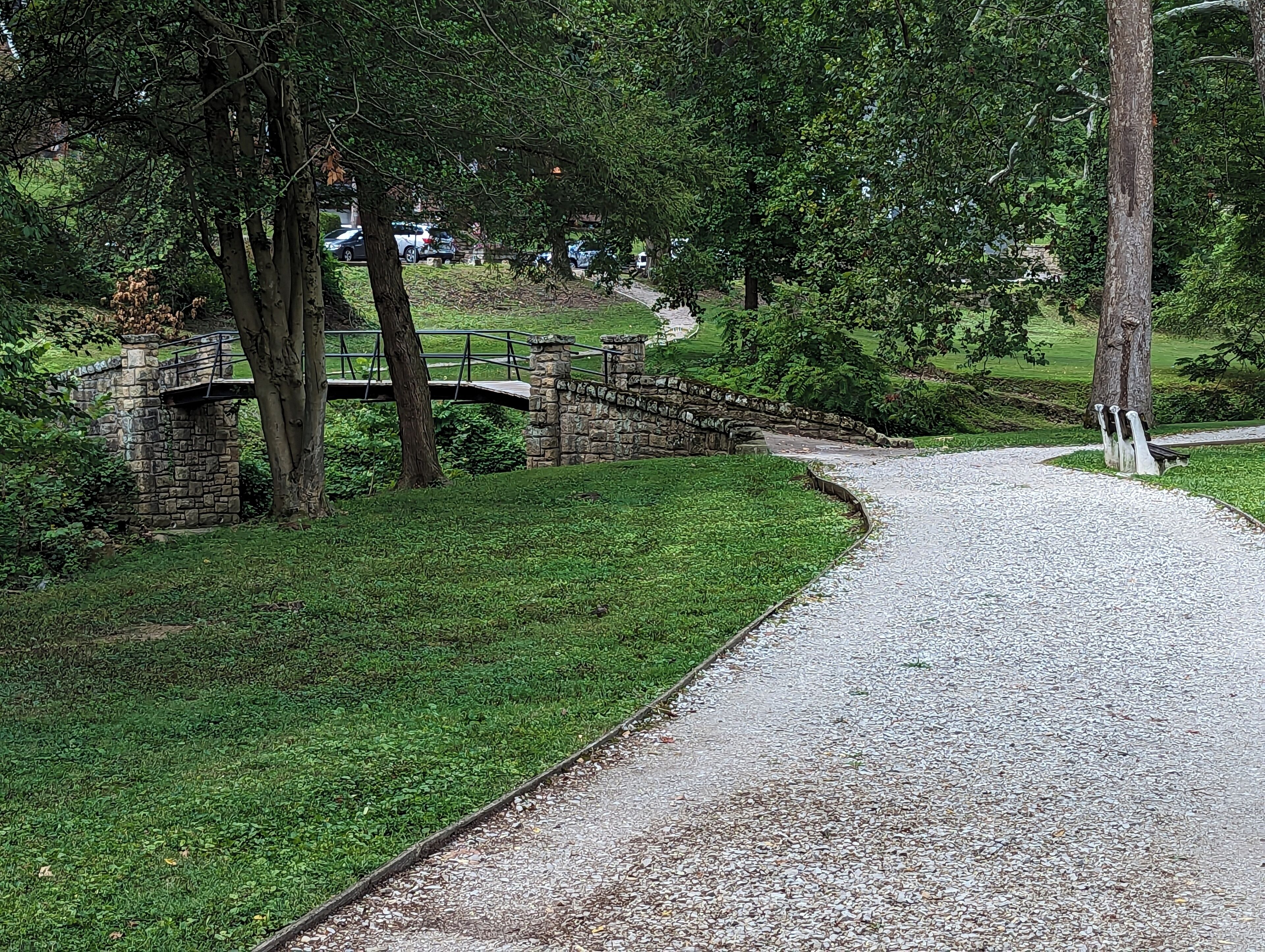 A gravel path leading to a stone bridge over a small stream, surrounded by lush greenery and trees.