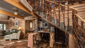 A rustic cabin interior with a wooden staircase, a leather sofa, a dining table, and a wooden desk. - Magical Treehouse on the Brazos River with spiral slide off the deck! 350 acres!  Tubing! (Weatherford)