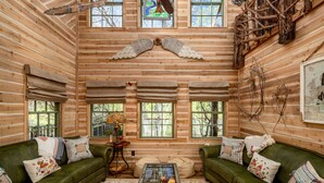 A cozy log cabin interior with two green sofas, a glass coffee table, and a wooden ceiling with a decorative angel wing. - Magical Treehouse on the Brazos River with spiral slide off the deck! 350 acres!  Tubing! (Weatherford)