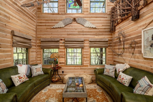 A cozy log cabin interior with two green sofas, a glass coffee table, and a wooden ceiling with a decorative angel wing.