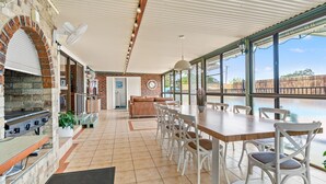 A spacious patio with a brick fireplace, a dining table with chairs, and large windows offering a view of a swimming pool. - Deco Beach - 9 Cedar Street (Evans Head)