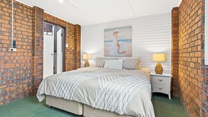 A bedroom with a large bed, two bedside tables with lamps, a painting on the wall, and brick walls. - Deco Beach - 9 Cedar Street (Evans Head)
