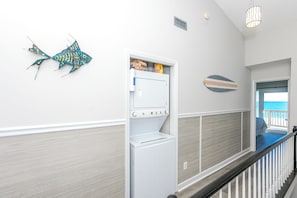 Corredor com parede branca, decoração em formato de peixe, máquina de lavar roupa e vista para o oceano através de uma porta aberta.