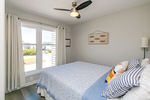 Een slaapkamer met een bed, gordijnen, een plafondventilator, een raam en een wanddecoratie.