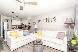 A modern living room with a white sofa, a small side table, a lamp, and a kitchen in the background.