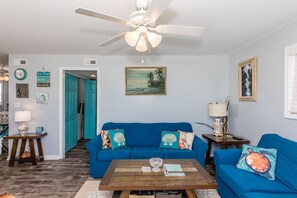 Un salon avec un canapé bleu, une table basse en bois, un ventilateur de plafond et un tableau accroché au mur.
