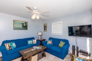 A living room with two blue sofas, a wooden coffee table, a ceiling fan, a flat-screen TV, and wall-mounted artwork. - Duzzi's Destiny - Oceanfront 5 BR Cottage!  Dog Friendly! (Surf City)