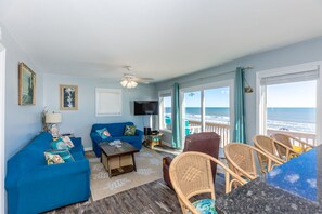 A living room with a blue sofa, a coffee table, and a TV. There are wicker chairs and a view of the ocean through large windows. - Duzzi's Destiny - Oceanfront 5 BR Cottage!  Dog Friendly! (Surf City)