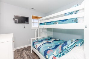 Un lit superposé avec une parure de lit aux motifs bleus et blancs sur le thème de l’océan, une commode blanche et un téléviseur fixé au mur.