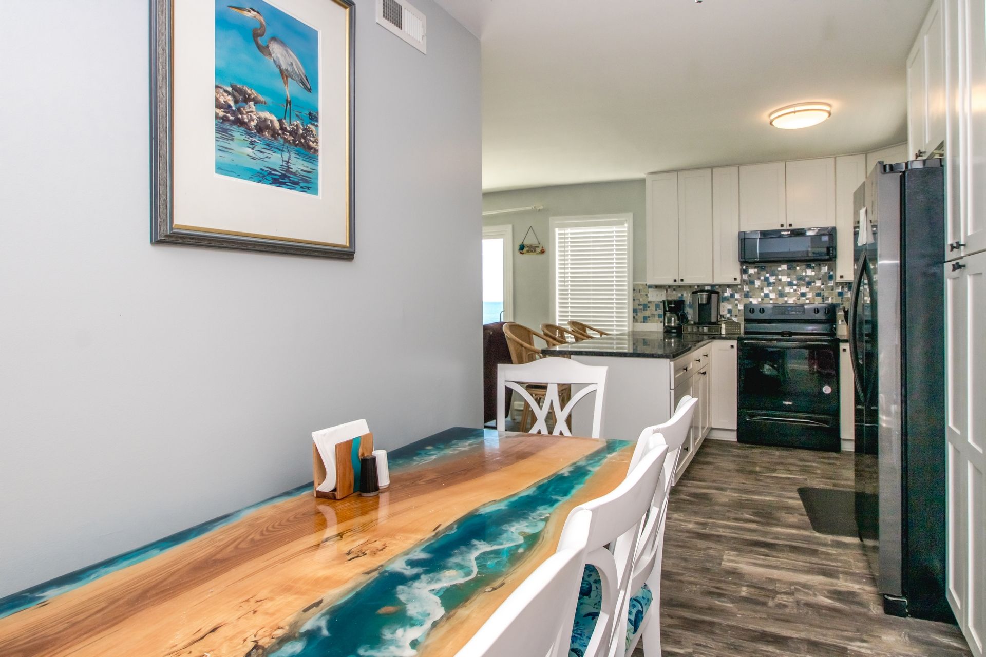 A dining area with a wooden table and chairs, a kitchen with white cabinets and appliances, and a framed picture on the wall.