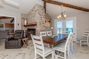 A dining area with a brick fireplace, white chairs, a wooden table, and a view of the ocean through a glass door. - Sandy Toes - Enormous 5BR Oceanfront Home with Apartment (Surf City)