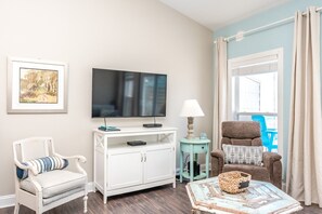 A living room with a television, white furniture, a blue chair, a lamp, and a window with curtains.