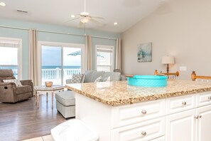 A spacious living area with a large window overlooking the ocean, a ceiling fan, and a kitchen island with a blue bowl.