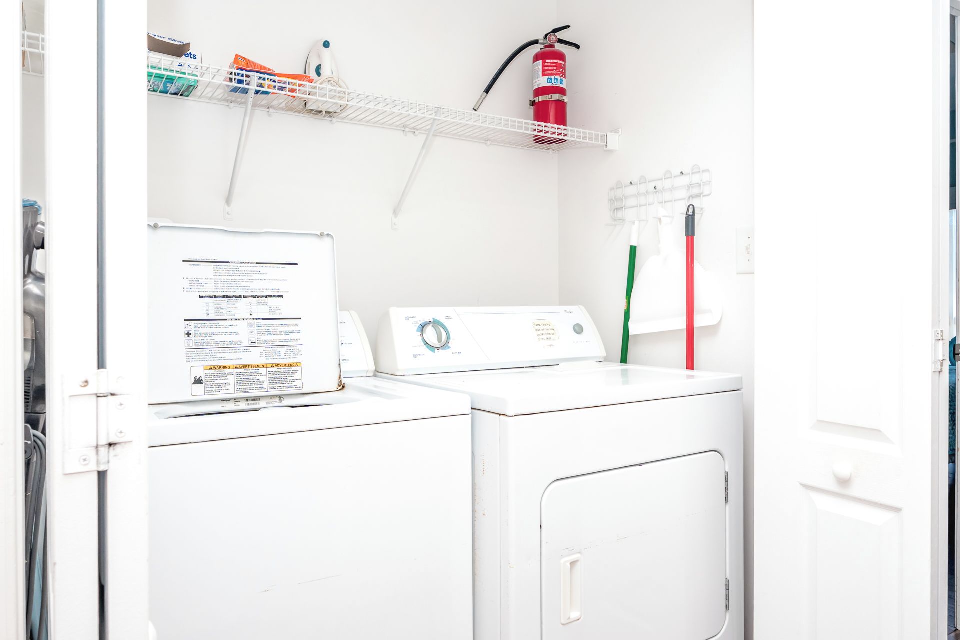 Een wasruimte met een witte wasmachine, een droger, een brandblusser en een plank met schoonmaakmiddelen.
