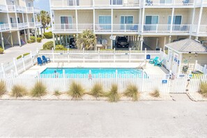 Un hôtel à plusieurs étages doté d’une piscine entourée d’une clôture en bois peinte en blanc, avec une plage au premier plan et un parking où sont stationnés des véhicules.