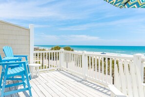 A wooden deck with a white railing, blue chairs, and a view of the ocean.