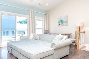 Un salón espacioso con un amplio sofá seccional gris, una mesa de madera y vistas al mar a través de amplias ventanas.