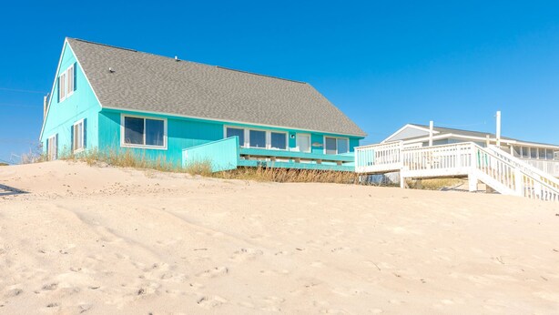 Ein Strandhaus mit steilem Dach, einer überdachten Veranda und einer Holztreppe, die zu einer Terrasse führt.