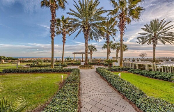 Un jardín arbolado con palmeras, un sendero pavimentado y vista al mar.
