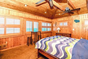 A room with wooden walls, a ceiling fan, a bed with a multicolored quilt, and a window with blinds.