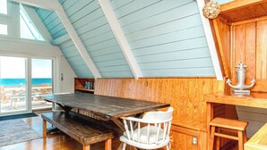 Ein Holzinnenraum mit hoher Decke, einem Esstisch, einem weißen Stuhl und Blick auf das Meer durch ein großes Fenster.