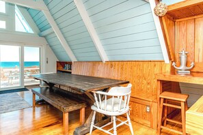 A wooden interior with a high ceiling, a dining table, a white chair, and a view of the ocean through a large window. - Barefoot Shack - Classic Custom 5BR Oceanfront Home - Great Views from Decks (Surf City)