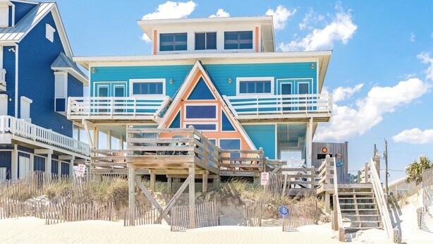 Ein mehrstöckiges Gebäude direkt am Strand mit Balkon, Holzstegen und einem sandigen Strand im Vordergrund.
