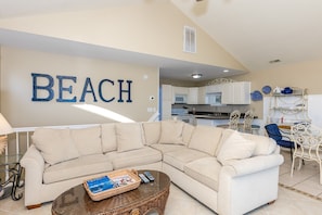 A living room with a large beige sectional sofa, a glass coffee table with magazines, and a kitchen in the background with white cabinets and a 'BEACH' sign. - Carolina Glory - 4BR Oceanfront Duplex with Covered Porch (Surf City)