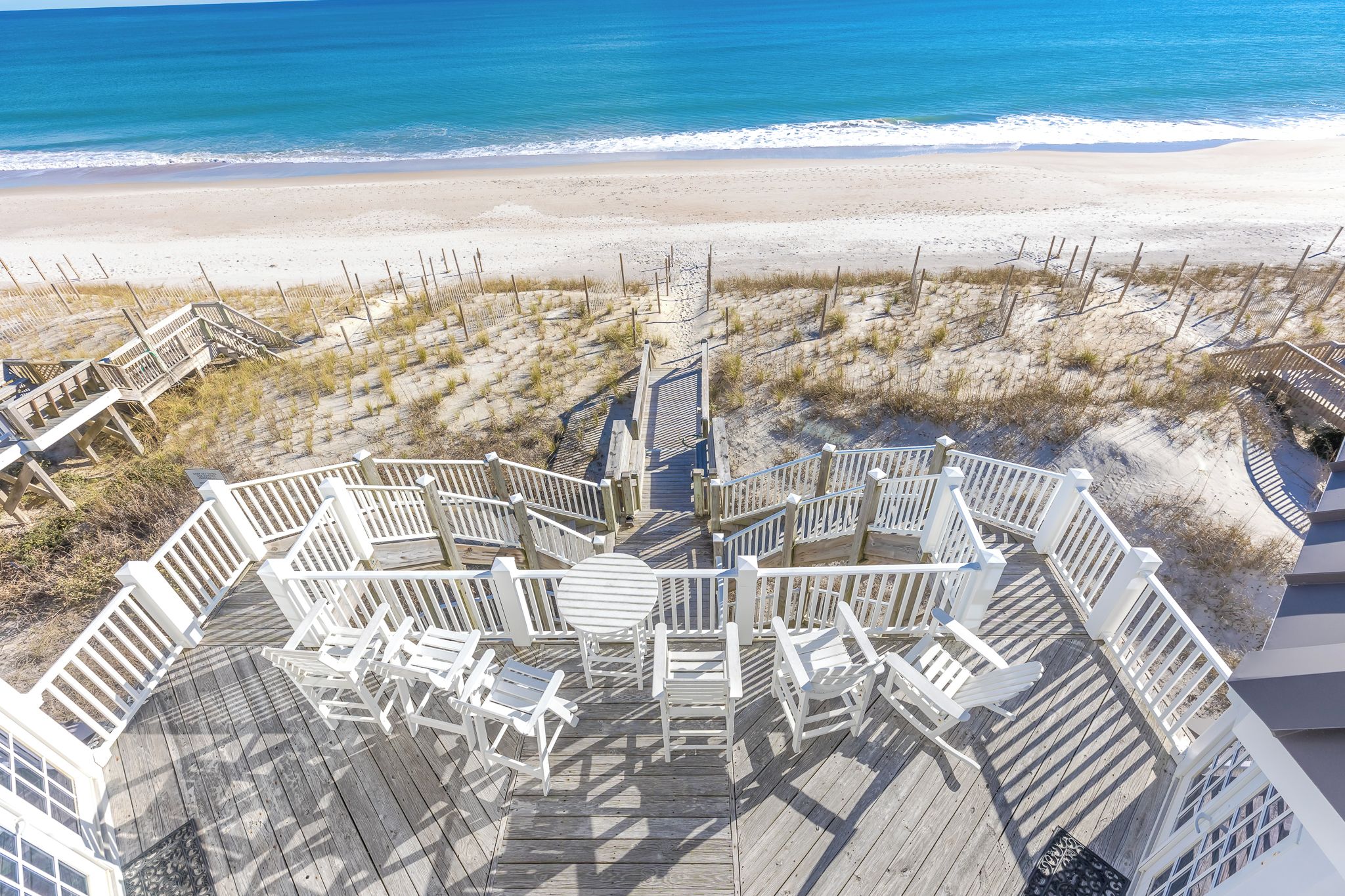 Eine Holzterrasse mit weißen Geländern und Stühlen mit Blick auf einen Strand.