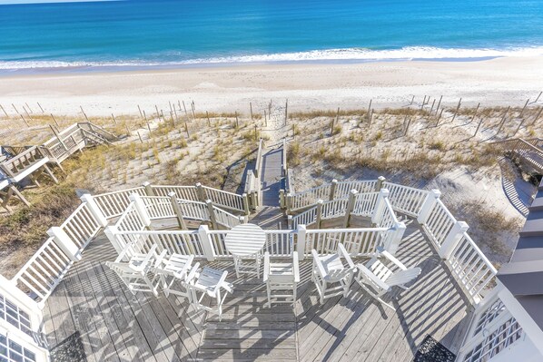 Une terrasse en bois, dotée de rampes et de chaises blanches, qui offre une vue sur la plage.