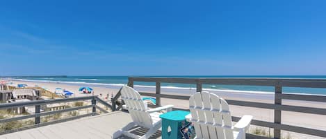 En terrasse ved stranden med to Adirondack-stole og et lille bord, med udsigt over en sandstrand og havet.
