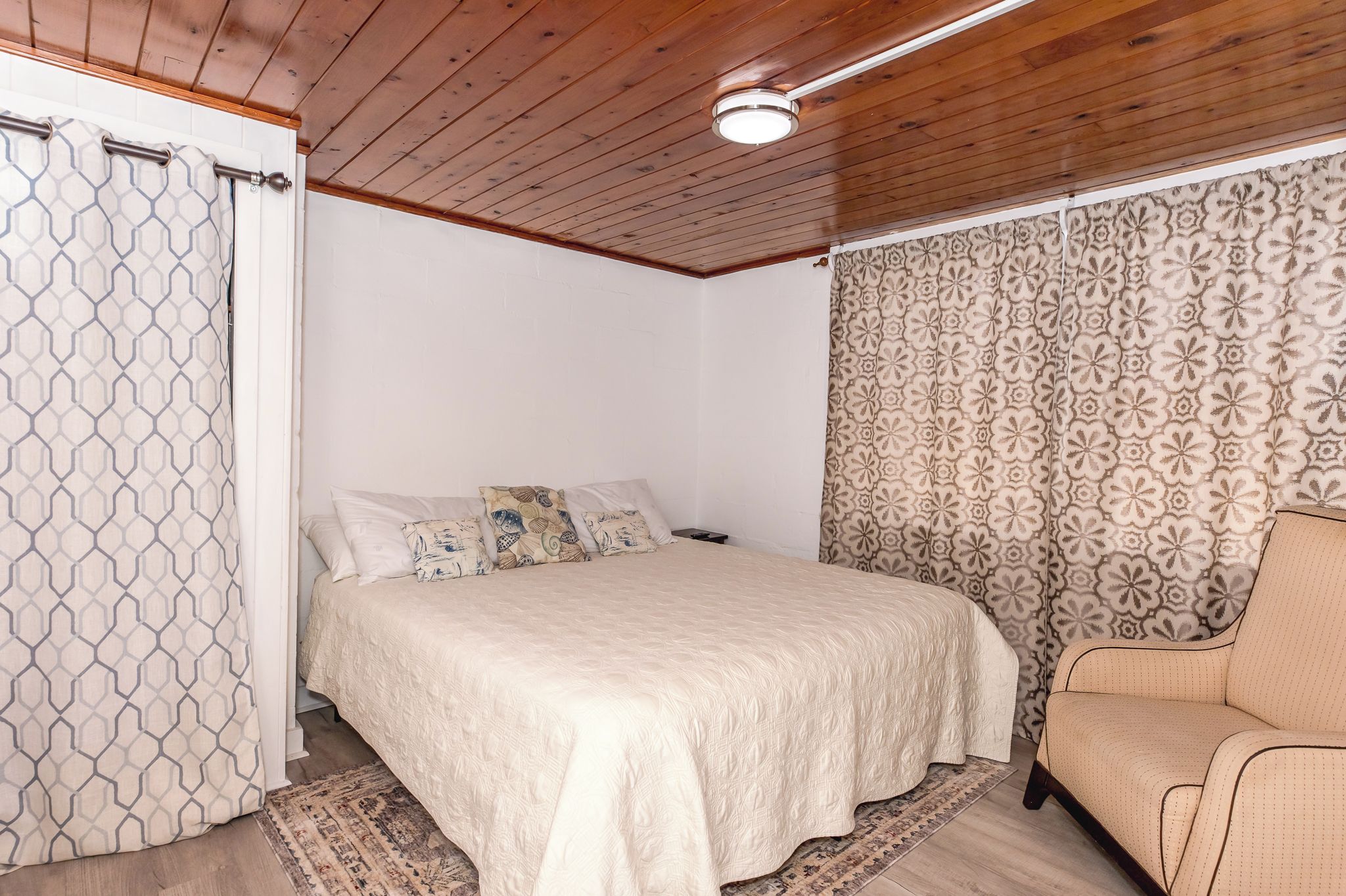 A bedroom with a bed, a chair, a patterned curtain, and a wooden ceiling.