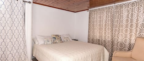 A bedroom with a bed, a chair, a patterned curtain, and a wooden ceiling.