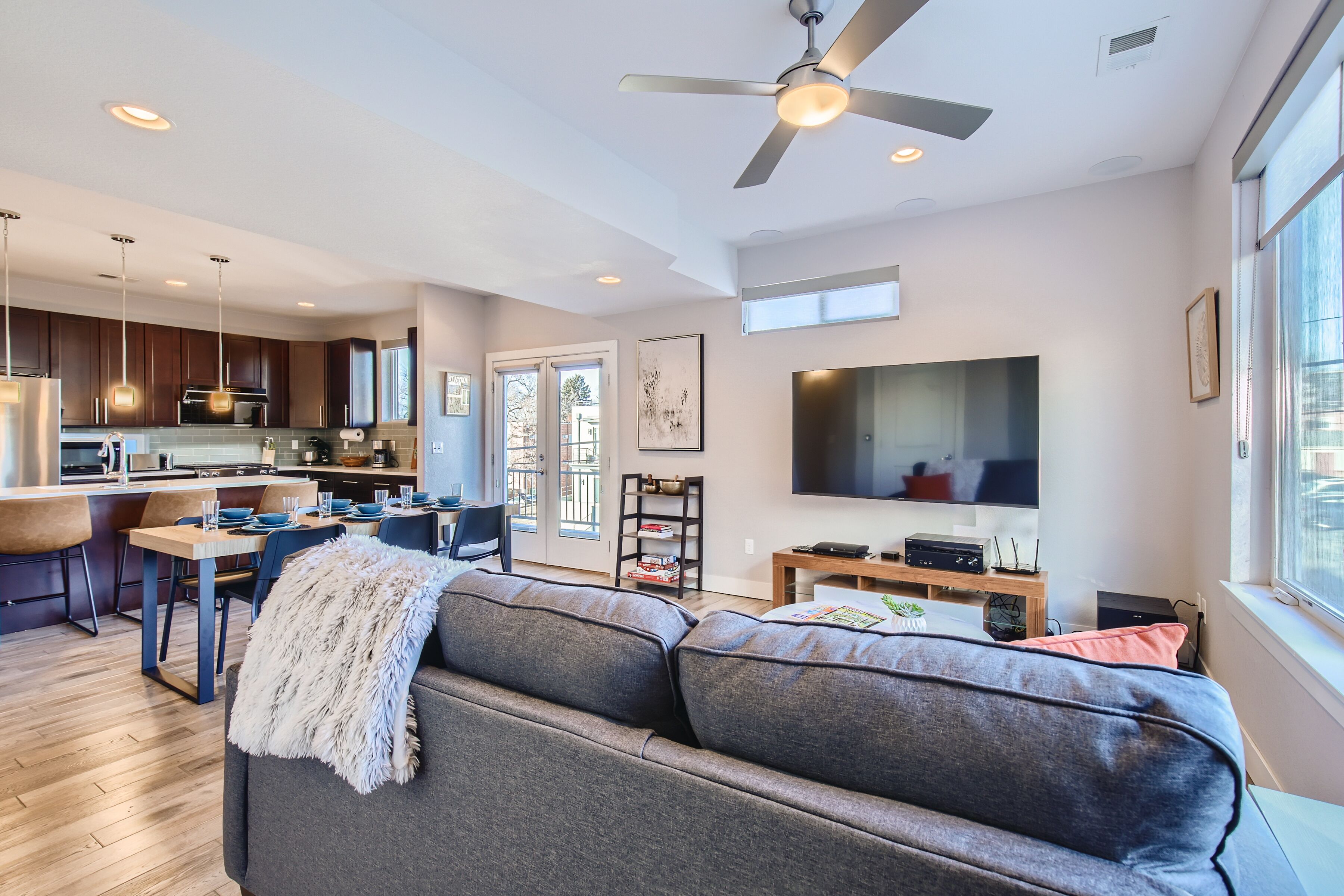 A modern kitchen with dark wooden cabinets, a dining area with a gray sofa, and a large flat-screen TV.