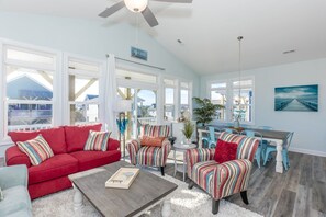 A spacious living area with a red sofa, striped armchairs, a dining table with chairs, and a view of the beach through large windows. - The Country Crab - 4BR Home, Elevator, Pet Friendly! (North Topsail Beach)