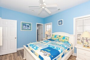 Una camera da letto con un letto, una ventola a soffitto, una porta, una finestra con persiane e un quadro appeso al muro.