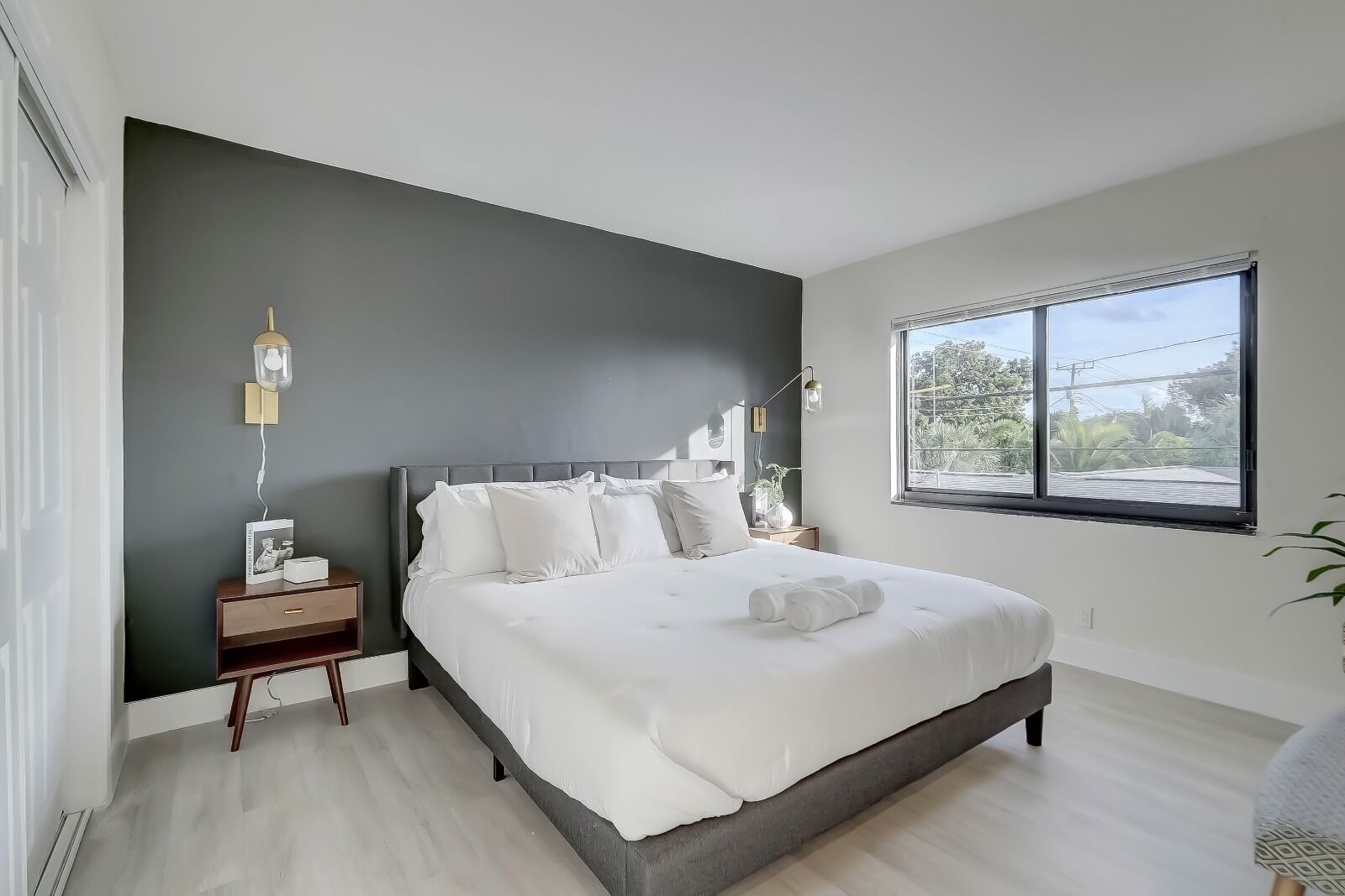 A modern bedroom with a large bed, a nightstand, a bedside table, a window, and a view of greenery outside.