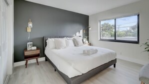 A modern bedroom with a large bed, a nightstand, a bedside table, a window, and a view of greenery outside. - Modern & Rlx space for 6 ppl (Fort Lauderdale)