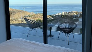 A balcony with a bed and two chairs, overlooking a coastal landscape with buildings and a road.