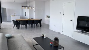 A modern living room with a sofa, a coffee table, and a TV. There is a dining area with a table and chairs in the background.