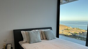 A bedroom with a large bed, a bedside table with a lamp, and a view of the ocean.