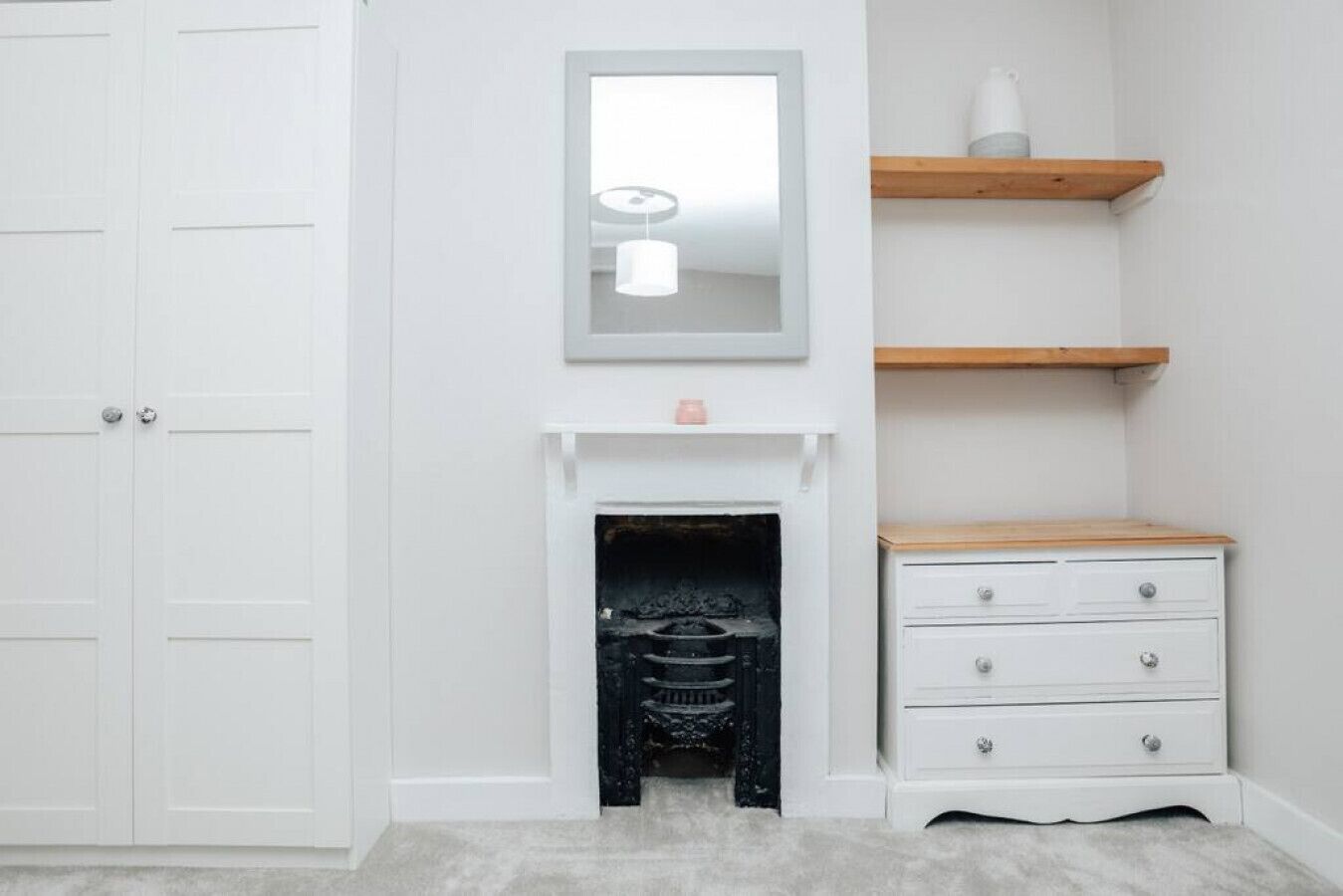 Um quarto claro com lareira, um espelho acima e uma prateleira de madeira com um vaso branco.