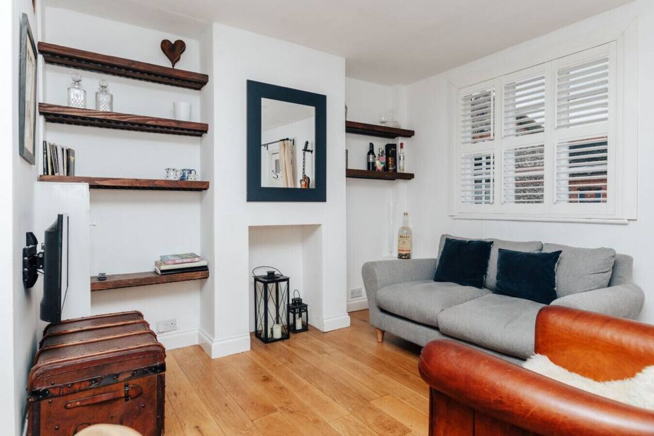 Un salon avec un canapé gris, des étagères en bois, un miroir et une fenêtre avec des stores.