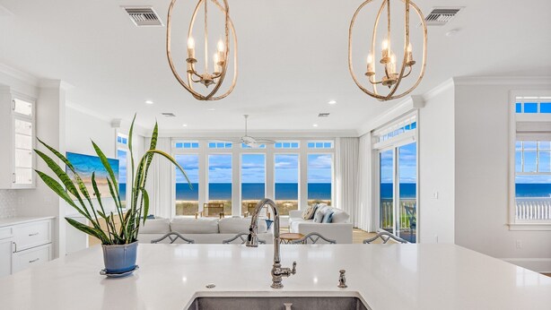 A modern kitchen with a large island, a sink, and a potted plant. There is a view of the ocean through large windows.