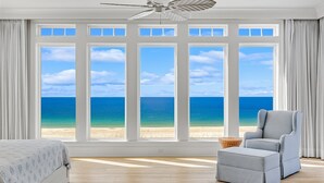 A bedroom with a large window, a ceiling fan, a bed, a chair, and a small ottoman.