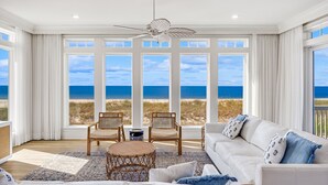 A spacious living room with a large window overlooking the beach, a ceiling fan, and a white sofa with blue pillows.