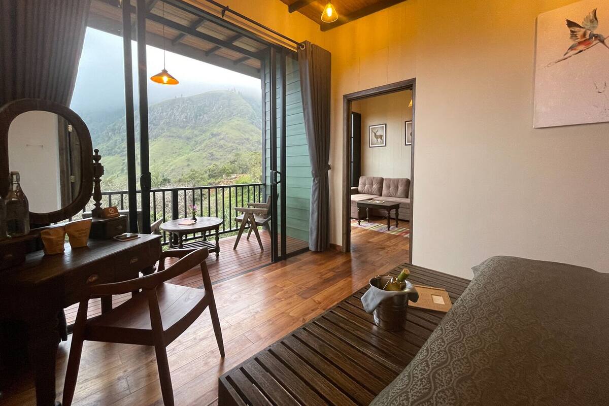 A room with a wooden floor, a dining table with chairs, a sofa, a small table with a bottle and a plant, and a large window with a view of green hills.