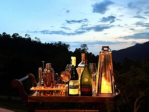 A table with various bottles and glasses, set outdoors with a mountainous backdrop. - The Capoe House (Kalupahana)